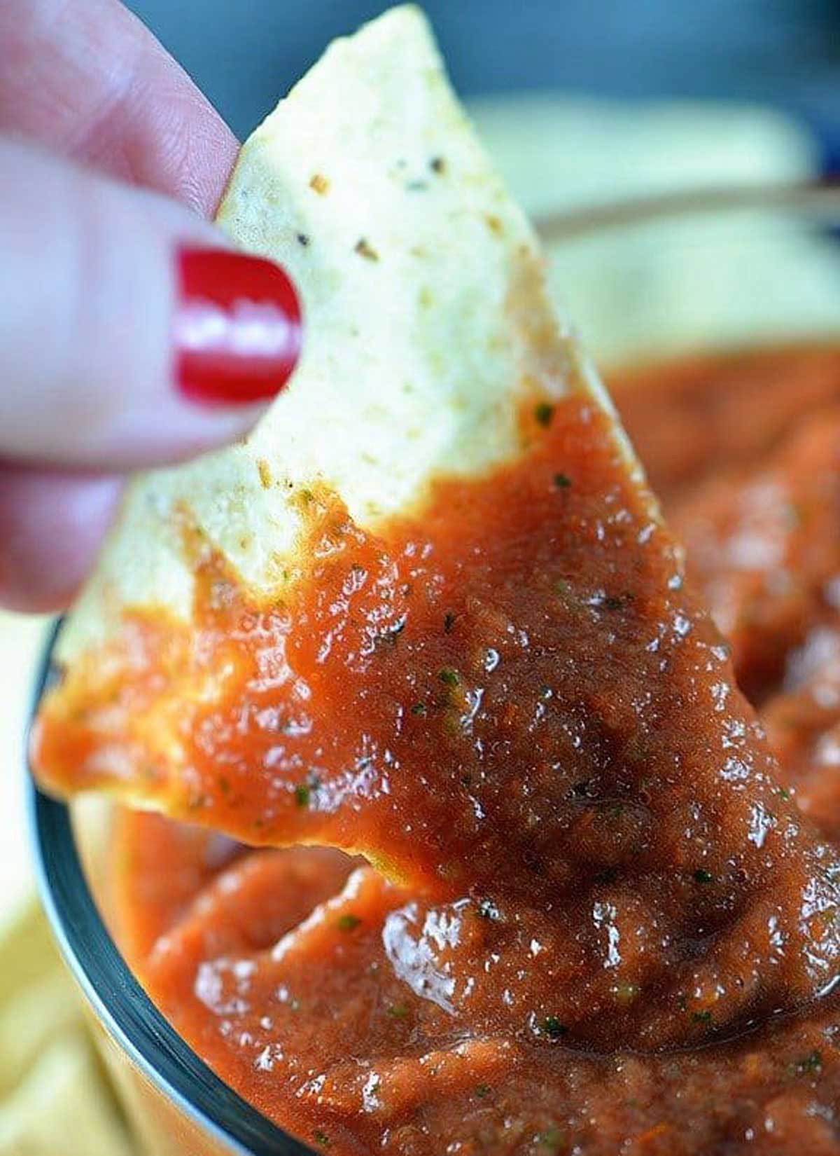 Glass container with salsa being dipped onto a tortilla chip held by a hand - addapinch.com