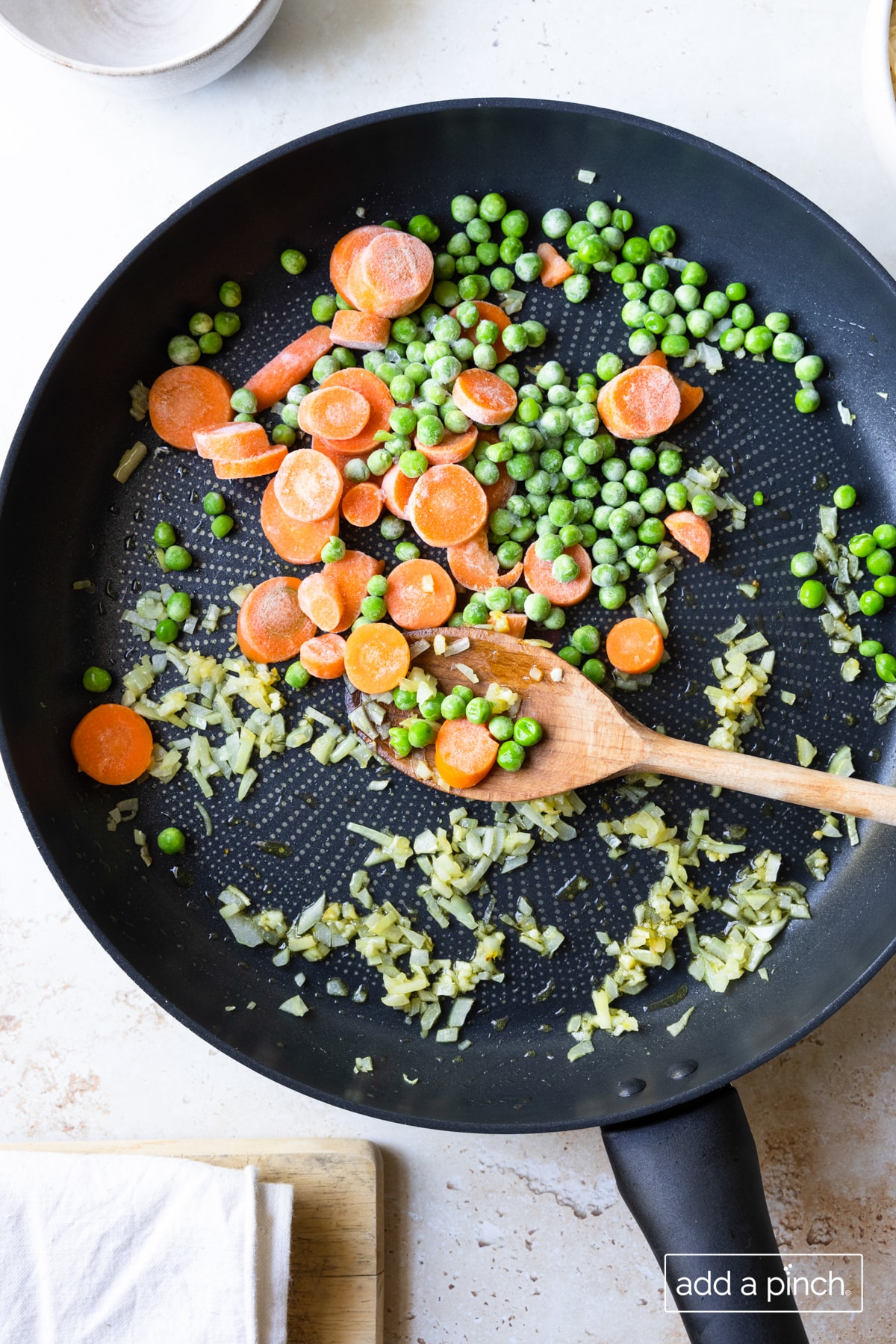 Photo of cooking garlic, onions, peas, and carrots in a skillet with a wooden spoon.