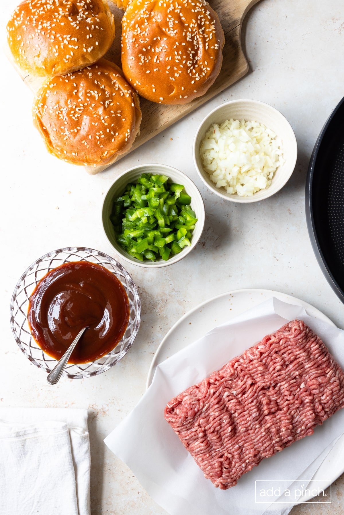 Photo of ground beef, onion, green pepper, bbq sauce, and buns on a white background.