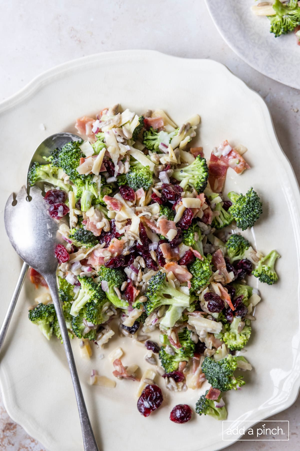 Broccoli salad on a white platter with serving utensils.