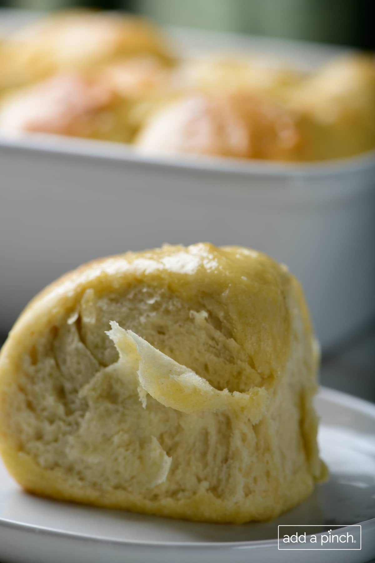 Photo of buttery, tender dinner roll on a white plate with baking dish in the background.