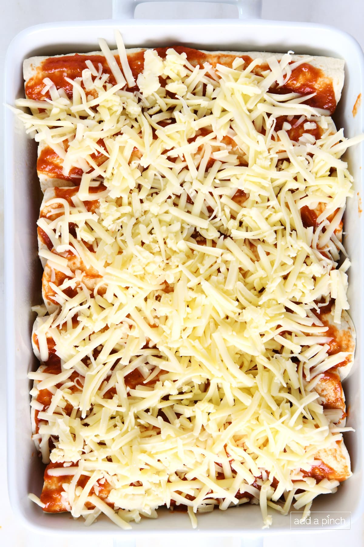 Chicken enchiladas topped with enchilada sauce and shredded cheese ready to be baked.