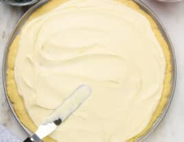 Photo of spreading cream cheese frosting on sugar cookie crust for fruit pizza with bowls of blueberries, strawberries, and cream cheese frosting.
