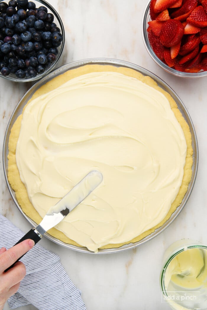Photo of spreading cream cheese frosting on sugar cookie crust for fruit pizza with bowls of blueberries, strawberries, and cream cheese frosting.