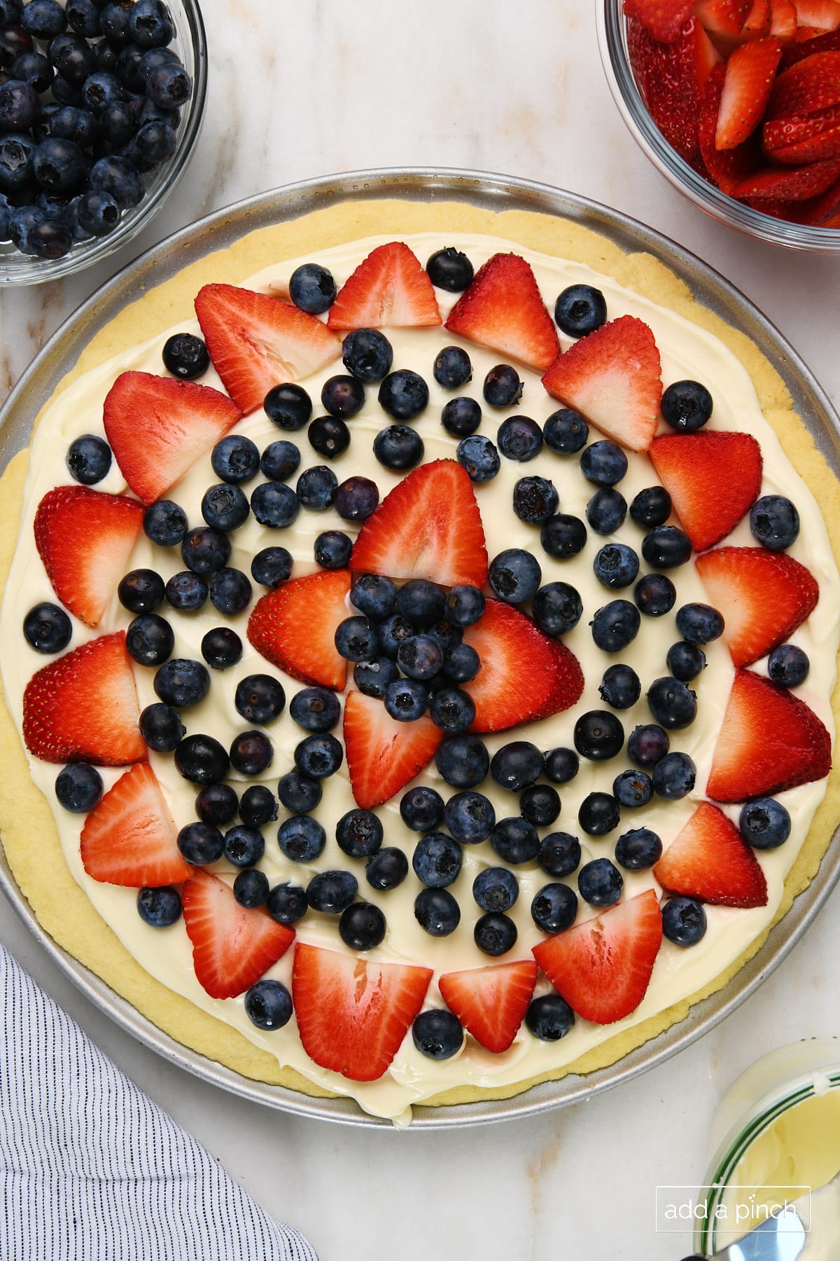 Fruit pizza on a marble surface surrounded by bowls of blueberries, strawberries, and cream cheese frosting.