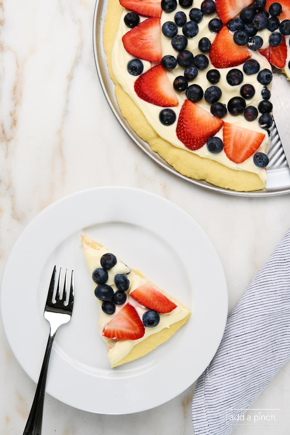 Slice of fruit pizza on a white plate with rest of fruit pizza to the side. 