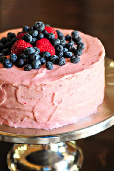 Strawberry cake with strawberry buttercream frosting topped with fresh blueberries and strawberries on a silver cake stand.