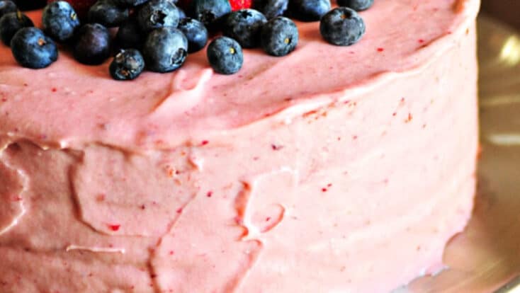 Strawberry cake with strawberry buttercream frosting topped with fresh blueberries and strawberries on a silver cake stand.