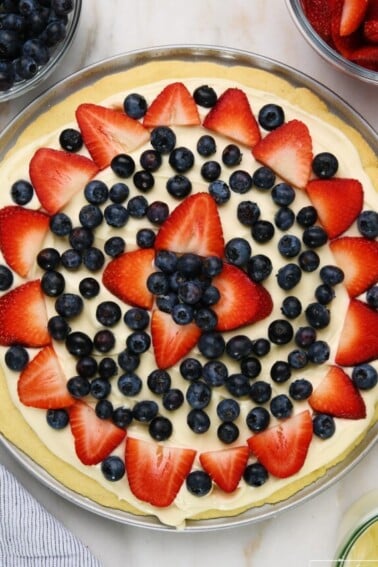 Fruit pizza on a marble surface surrounded by bowls of blueberries, strawberries, and cream cheese frosting.