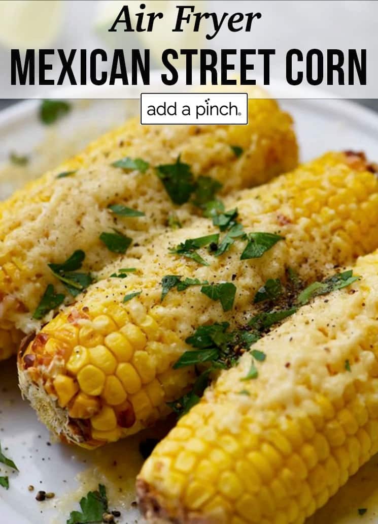 Three ears of Mexican Street Corn garnished with cheese, herbs on a white plate - with text - addapinch.com
