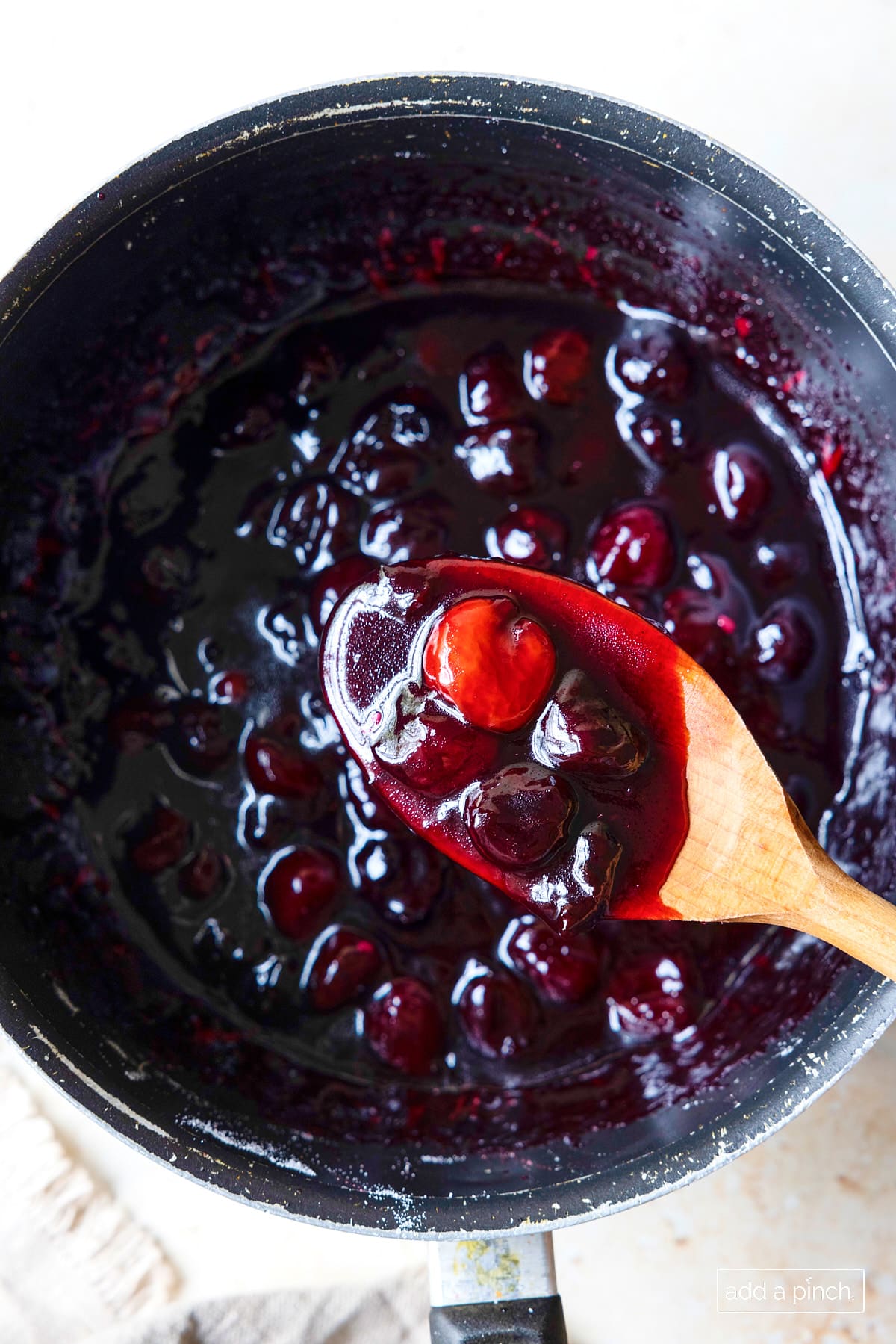 Homemade cherry pie filling in a sauce pan with a wooden spoon.