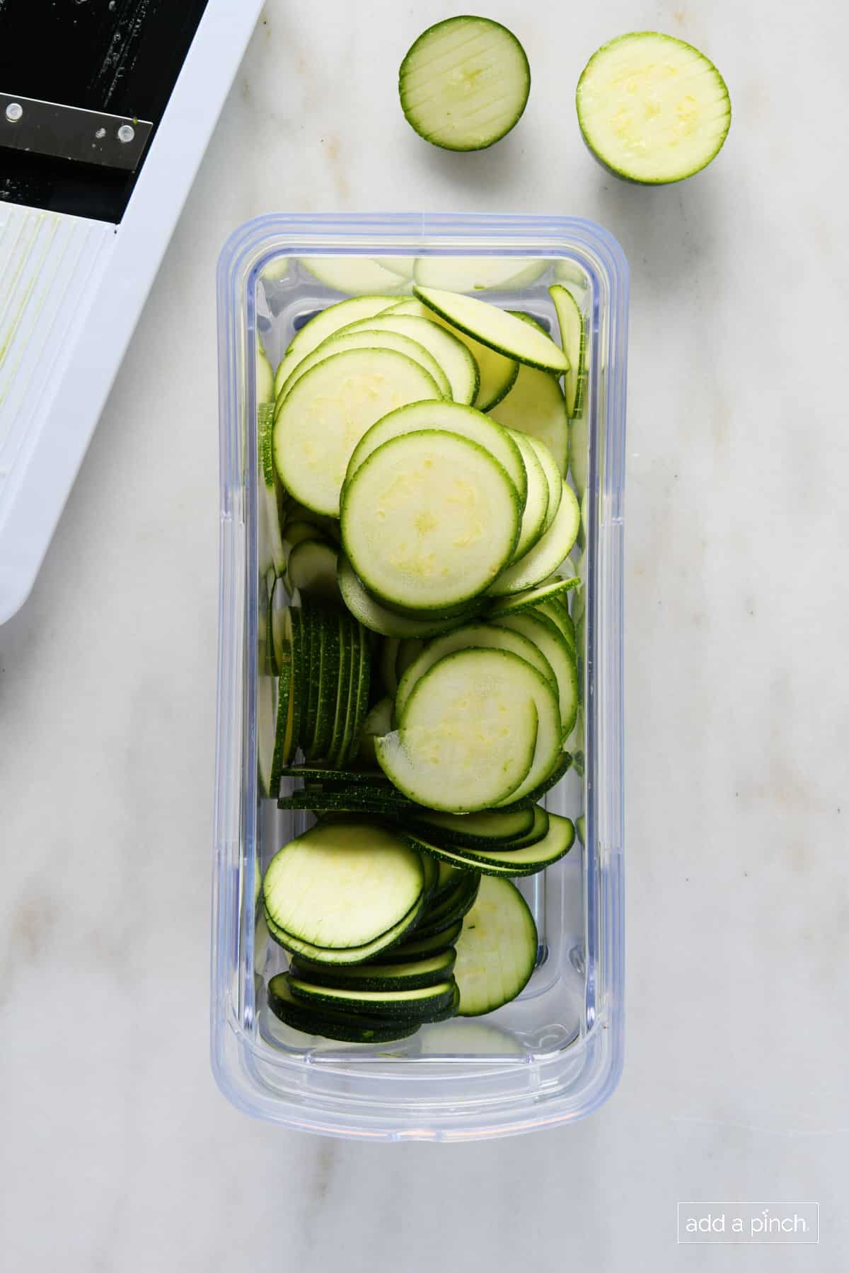 Mandoline slicer sits beside a big container of thinly sliced zucchini rounds on a marble counter // addapinch.com