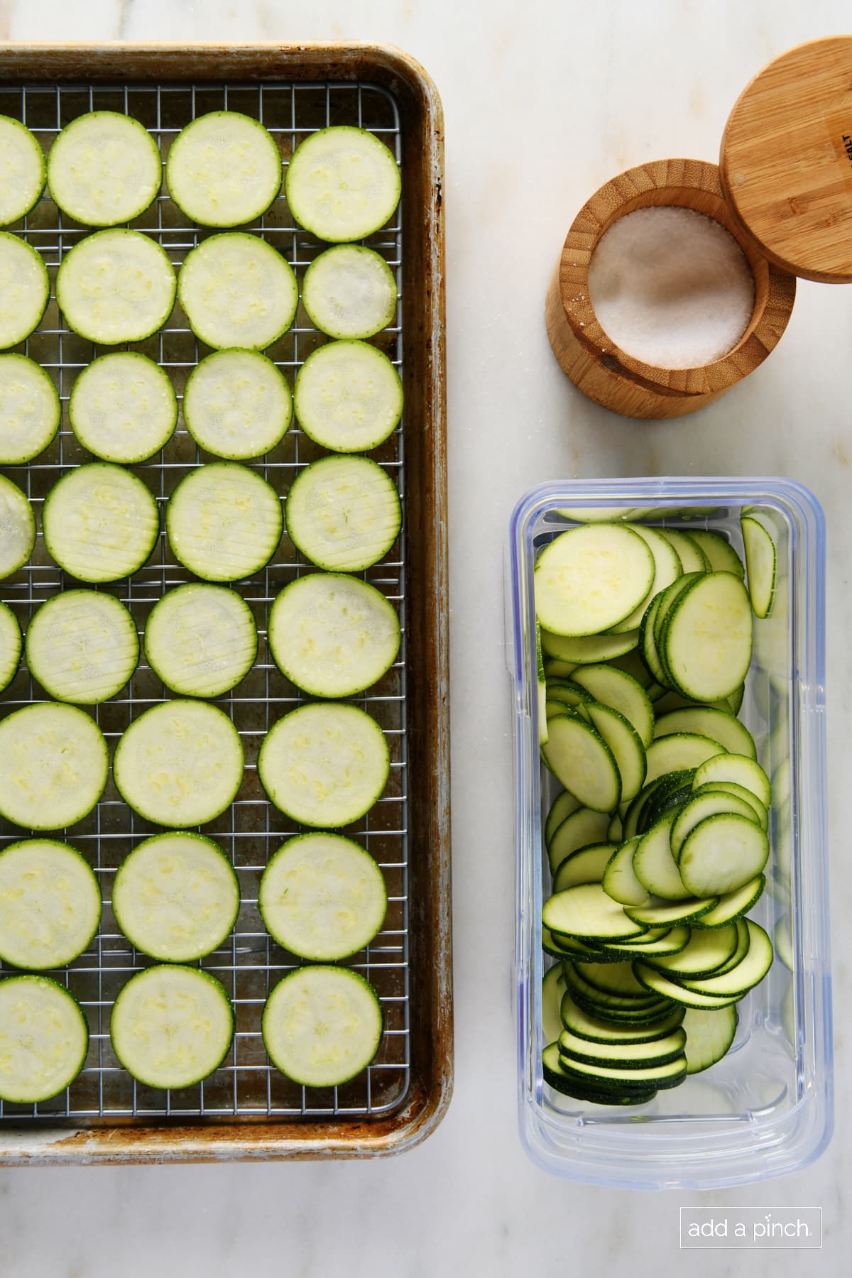 Wired rack in a rimmed baking sheet holds rows of zucchini crisps beside a wooden salt container and container of zucchini slices //addapinch.com