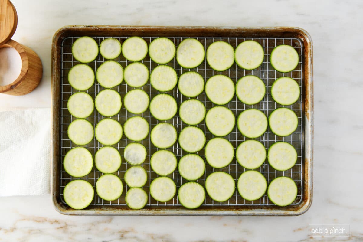 Overhead photo of baking sheet with rows of zucchini rounds beside a salt cellar and paper towel for blotting off excess moisture //addapinch.com