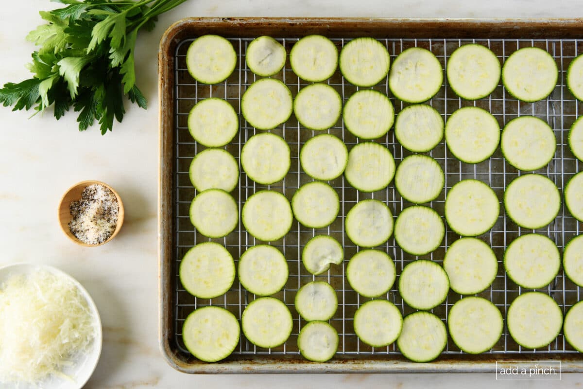Baking sheet holds zucchini slices beside bunch of fresh parsley, container of Stone House Seasoning and bowl holding grated Parmesan // addapinch.com