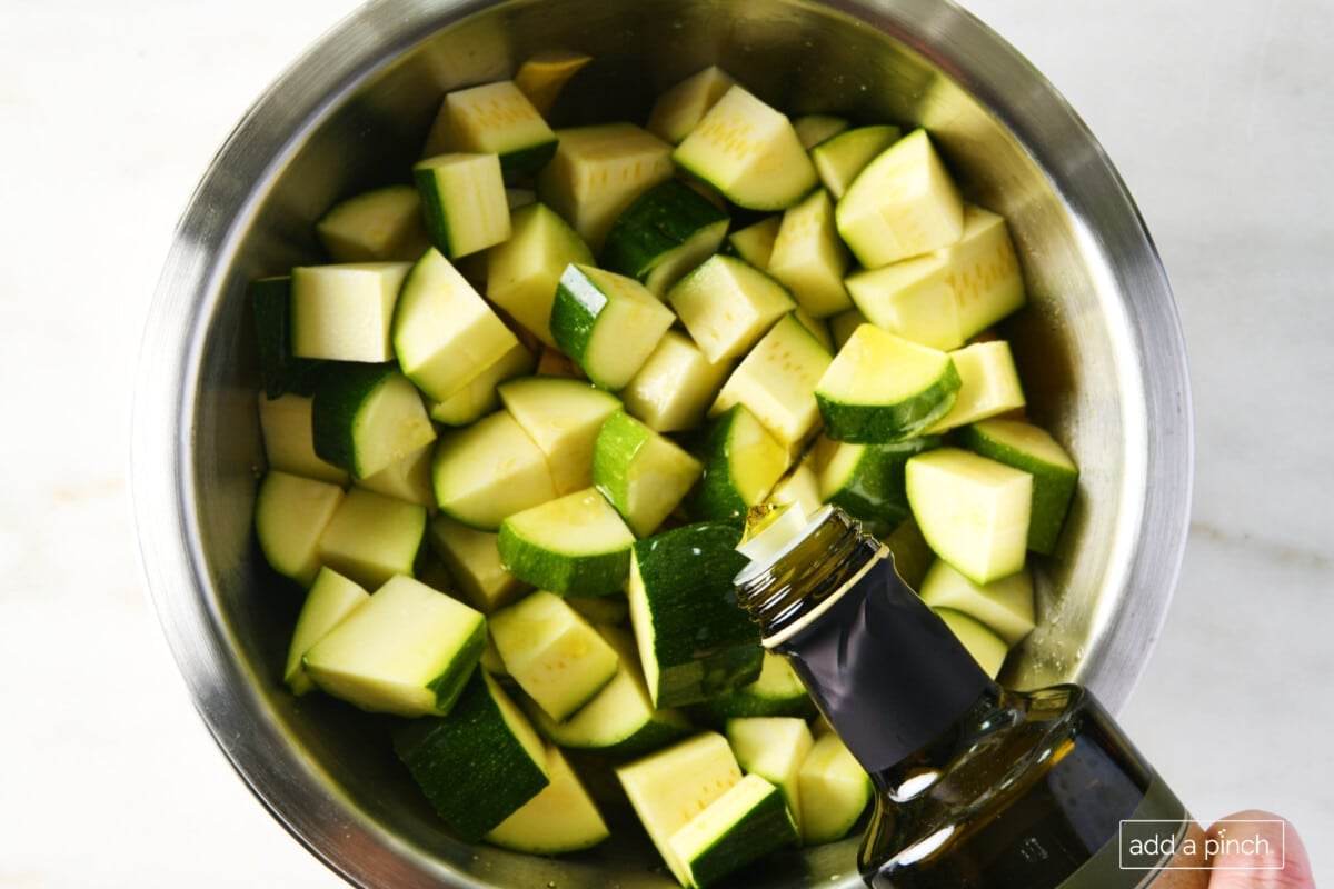Olive oil being poured into bowl of cut yellow squash and zucchini in a mixing bowl.