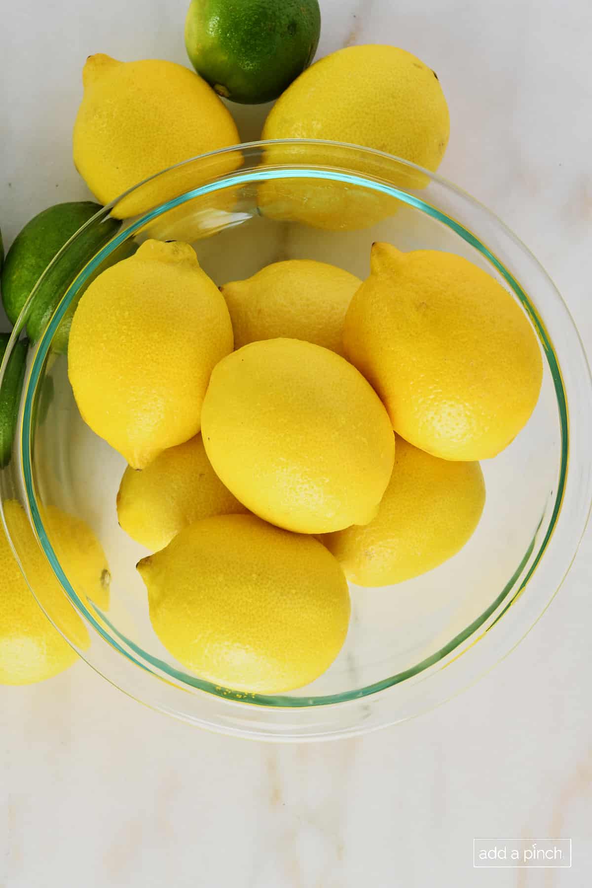glass mixing bowl filled with yellow lemons and a few limes on the side of the bowl set on a marble surface.