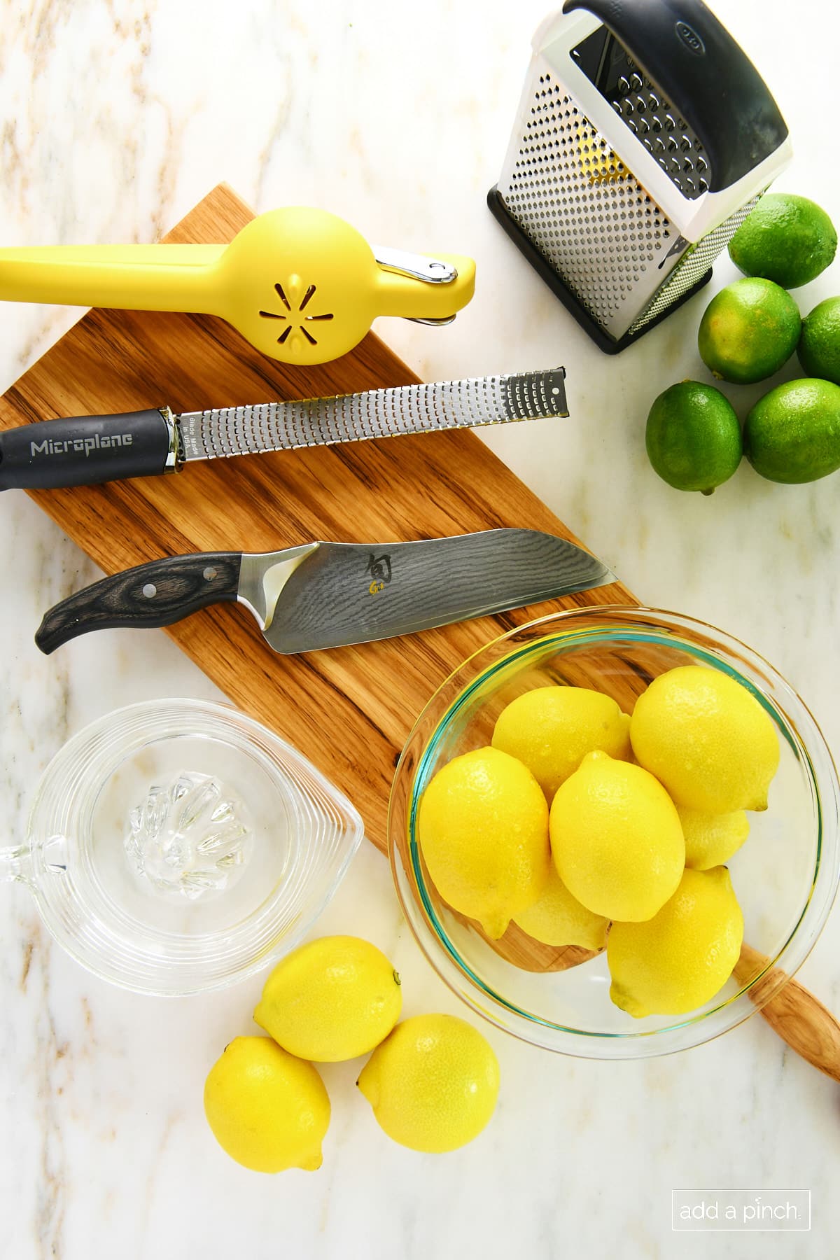 photo of marble surface with bowl of yellow lemons, chef's knife, yellow citrus juicer, and microplane on a cherry wooden board with yellow lemons and clear glass citrus juicer/reamer on left side of board and box grater and green limes on right side of board