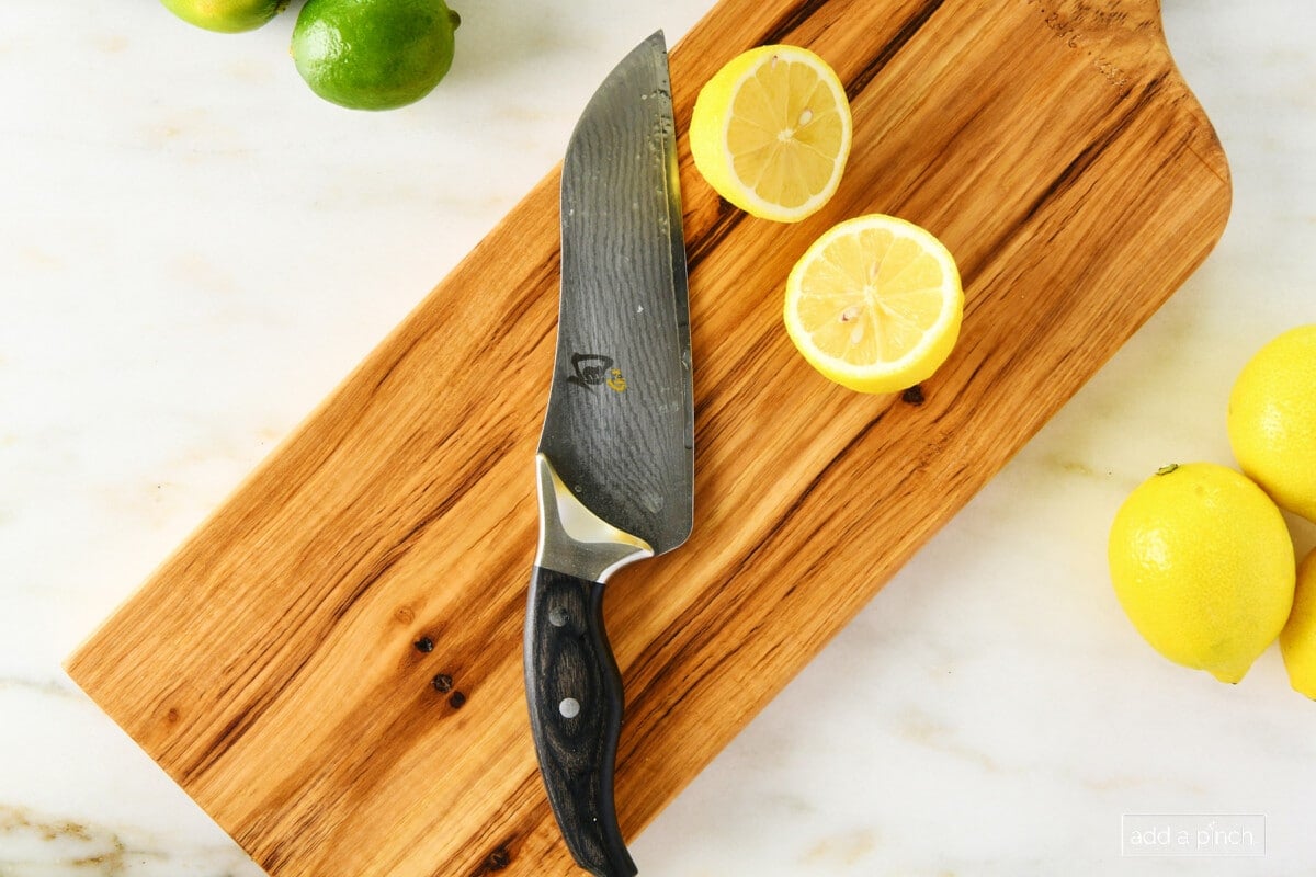 marble surface with chef's knife and yellow lemon cut in half on cherry wooden board with lemons on right side of board and green lime on top left side of board