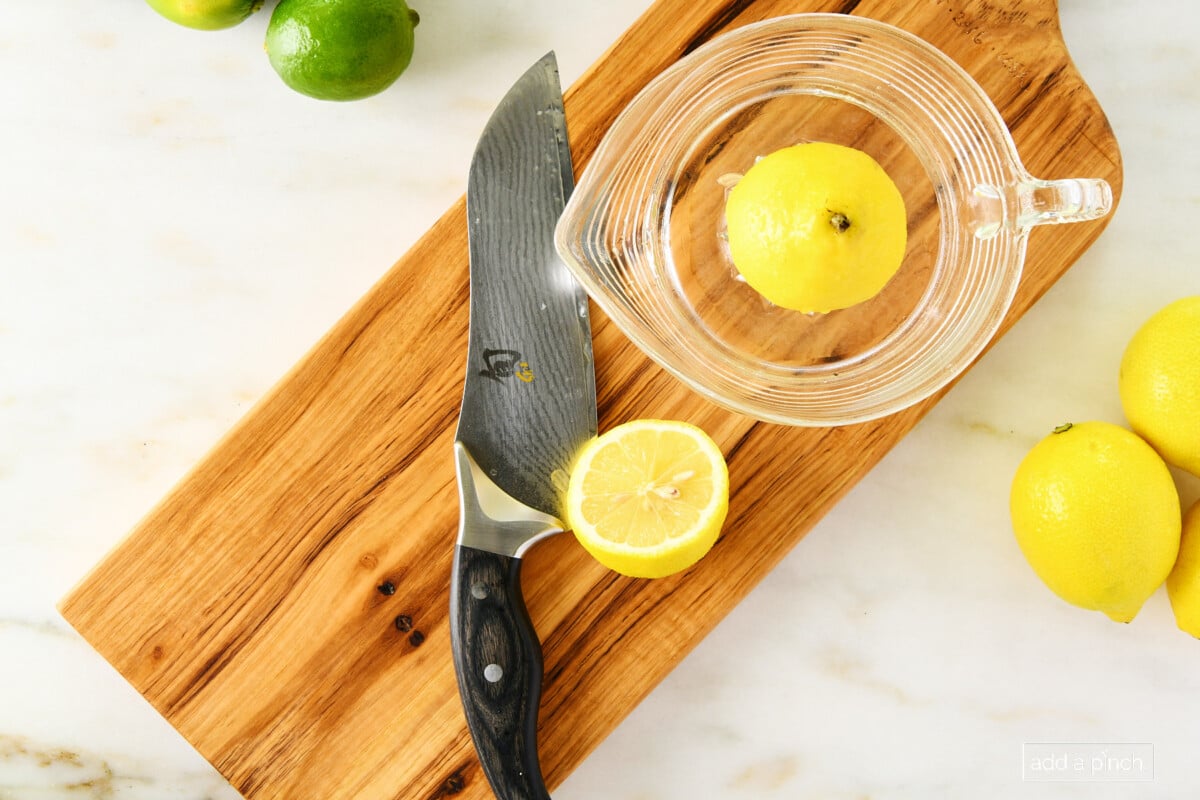 photo of cherry board with a chef's knife, yellow lemon half, and clear glass citrus reamer/juicer with a yellow half lemon on the reamer sitting on a marble surface and a green lime in the top left 