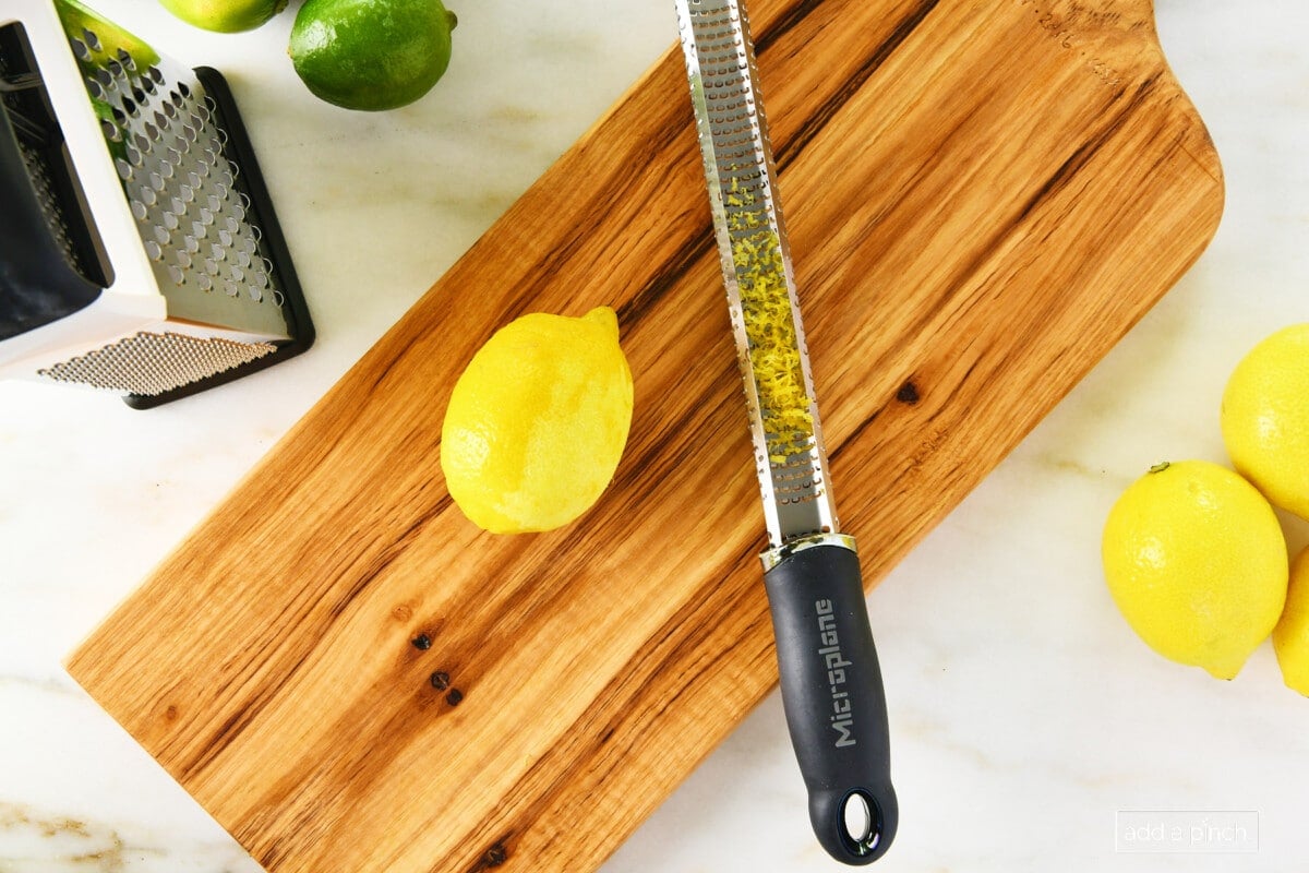 photo of cherry board with  microplane containing yellow zest and a yellow lemon on a marble surface with a yellow lemon on right side of board and a box grater and a green lime on the top left of the board
