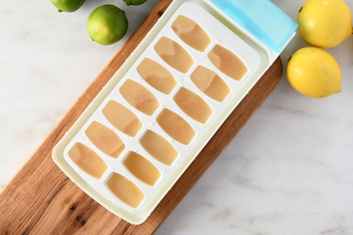 photo of white ice tray with frozen lemon juice sitting on cherry wooden board on marble surface with yellow lemons on top right of photo and green limes on top left