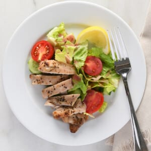 Sliced chicken thighs on a white plate with a garden salad and lemon wedge.