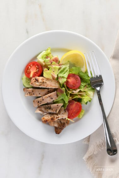 Sliced chicken thighs on a white plate with a garden salad and lemon wedge.