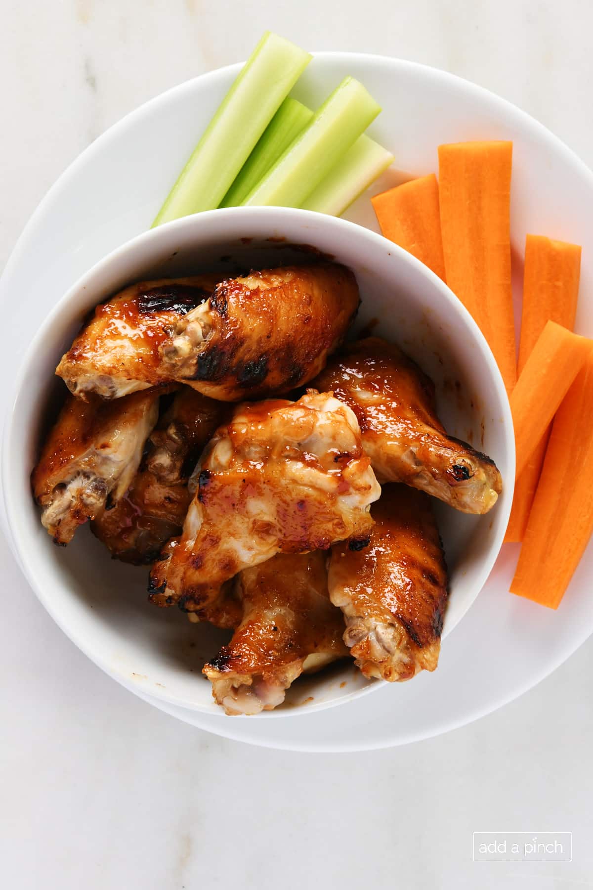 BBQ Chicken wings in a white bowl with carrots and celery on a marble surface.