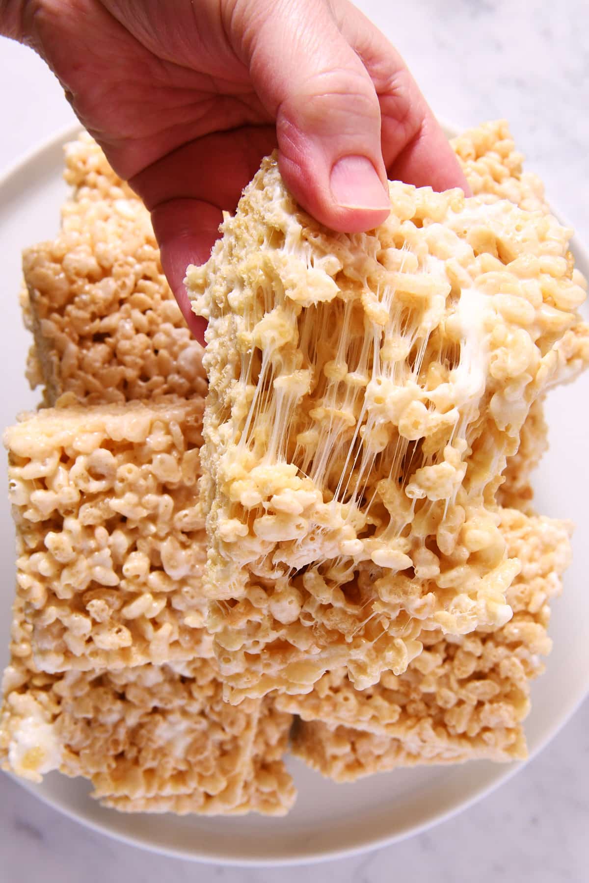Rice cereal treats stacked on a white cake plate with one treat pulled apart to show the gooey marshmallow.
