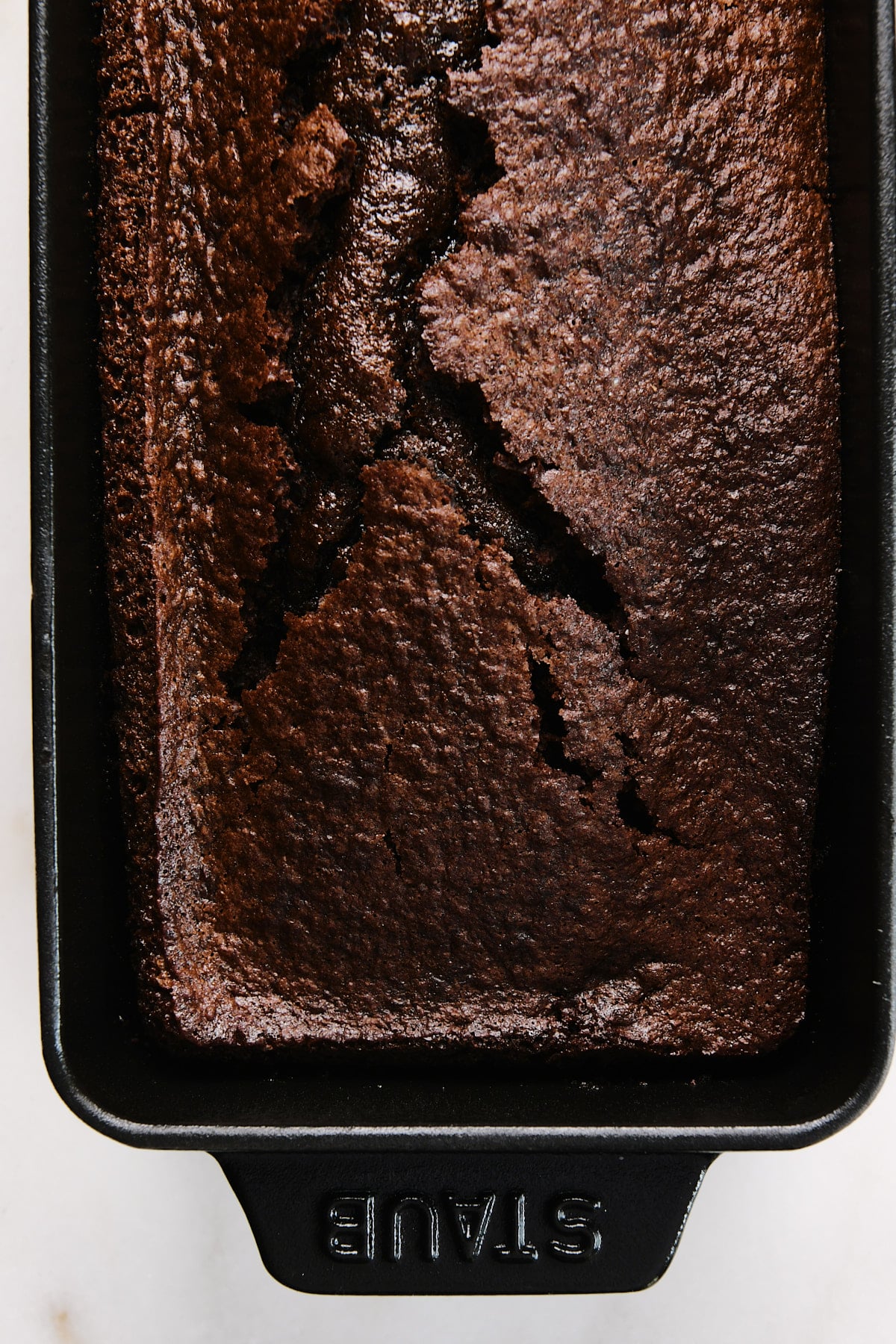 Chocolate loaf cake cooling in a loaf pan on a marble surface.