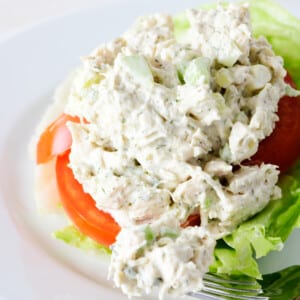 Classic chicken salad on a white plate with slice of tomato, lettuce, and a fork with a bite of chicken salad.