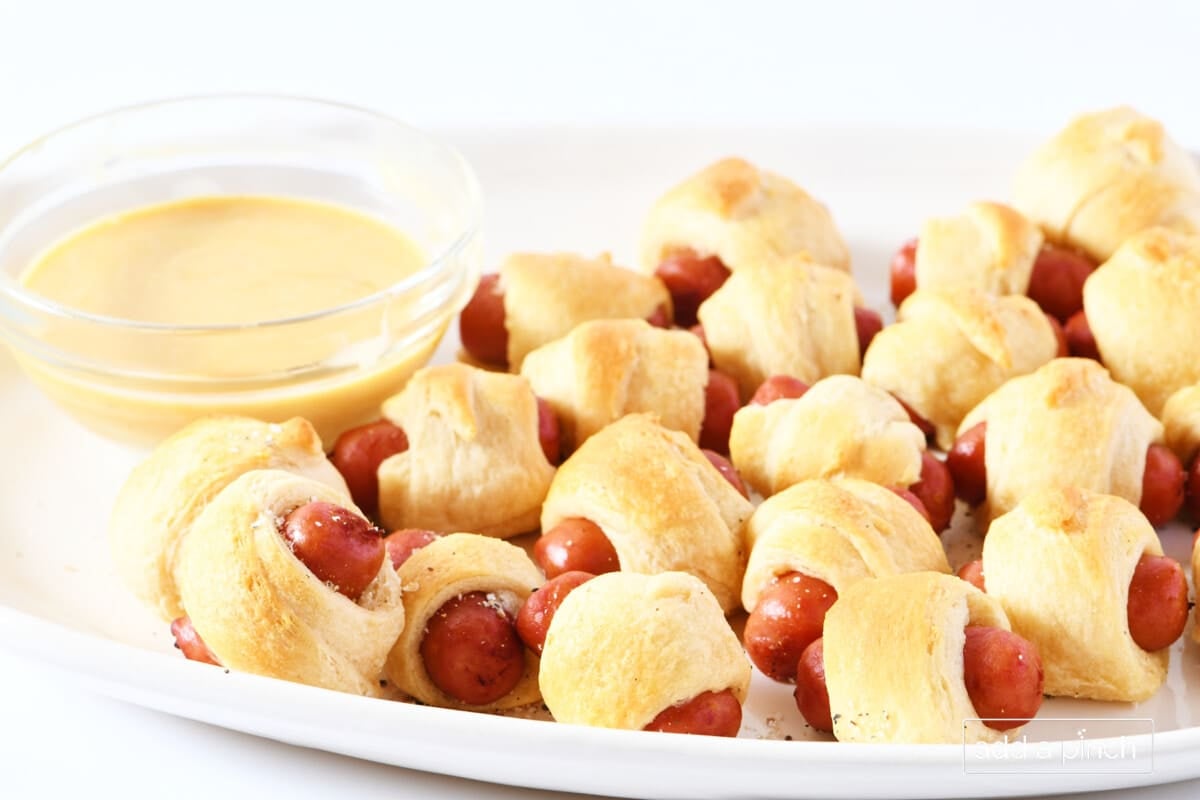 Pigs in a blanket on a white serving platter with dipping sauce in a glass bowl.