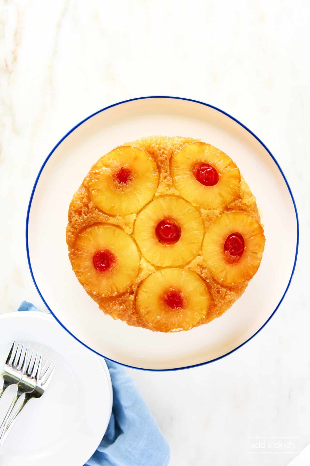 Homemade pineapple upside down cake on a white cake stand with a blue rim on a marble counter with stack of white plates and forks on a blue naplin.