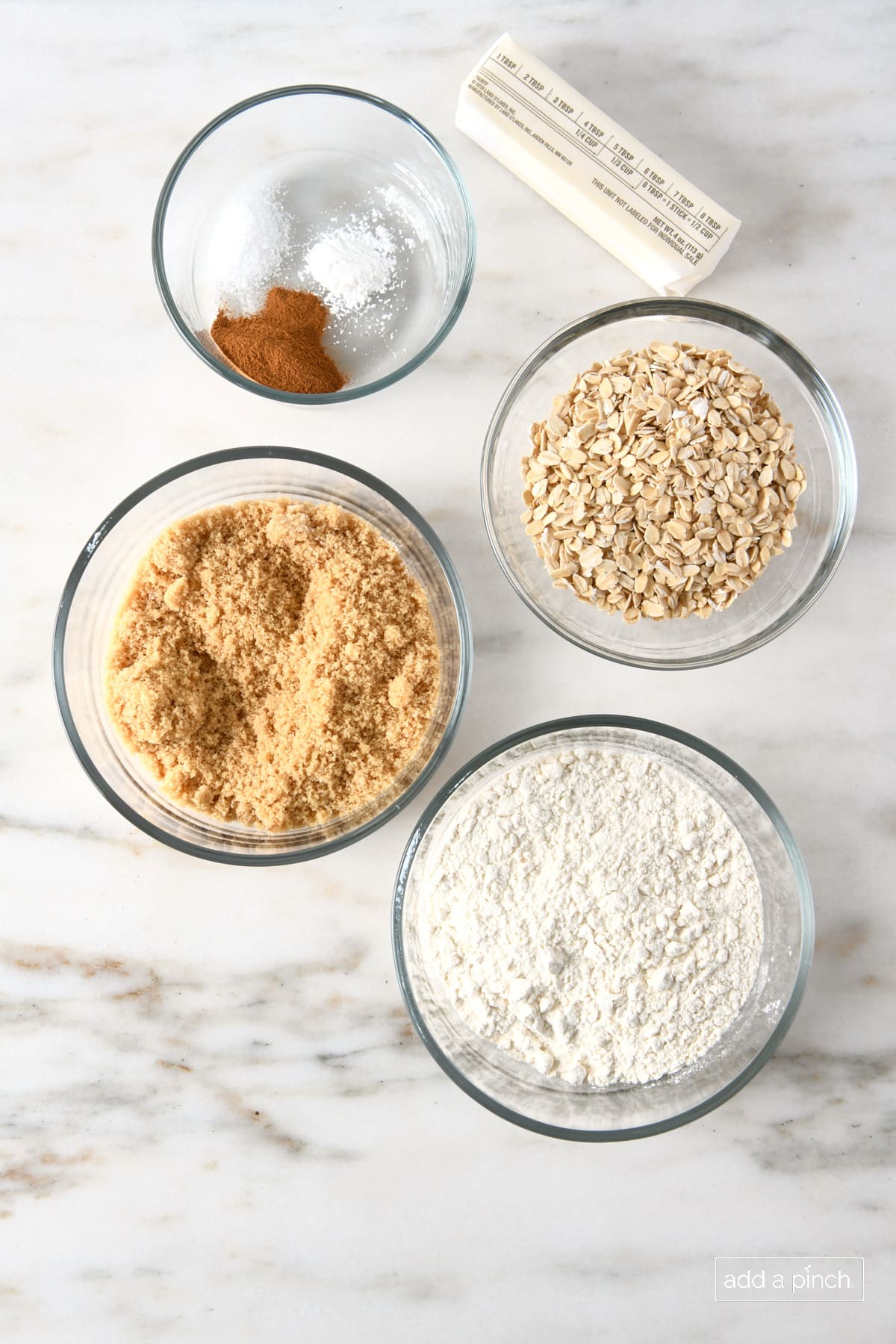 Flour, brown sugar, oats, salt, cinnamon, baking powder in glass bowls and butter on marble counter.