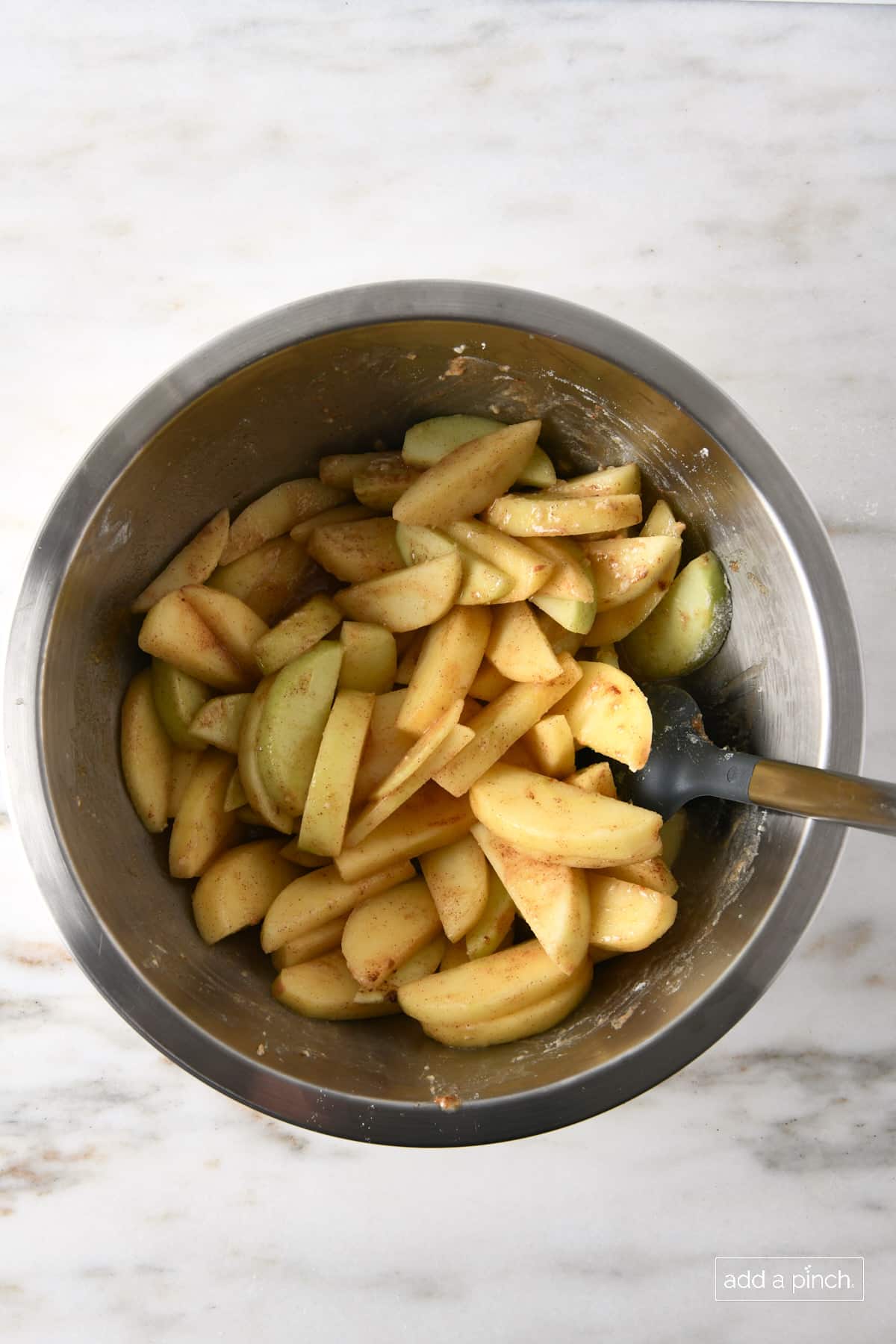 Stainless mixing bowl on marble counter holds apple slices coated in cinnamon, brown sugar and other apple filling ingredients.