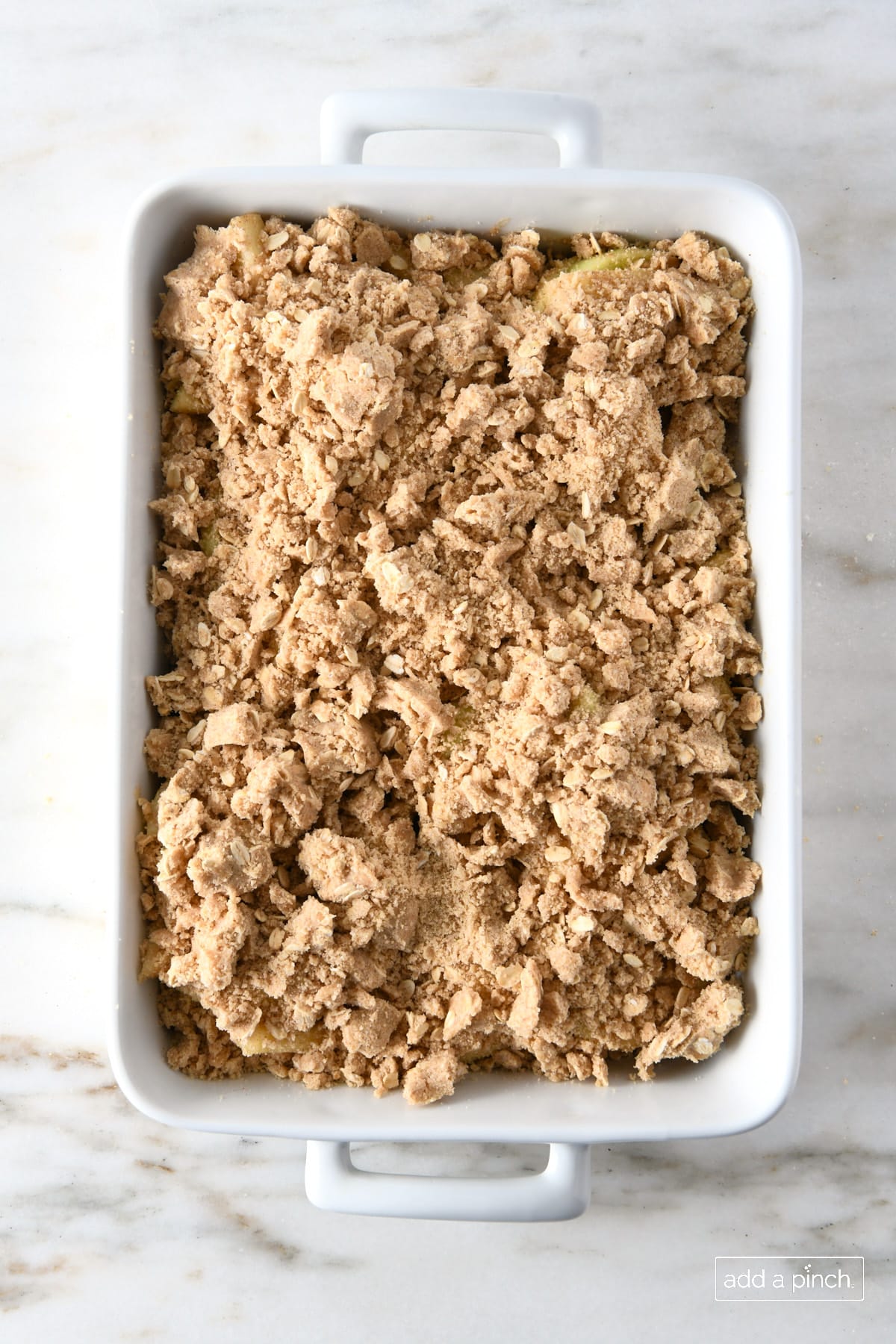 White baking dish on marble countertop holds unbaked apple crisp with apple slice peeking out of oat streusel topping.