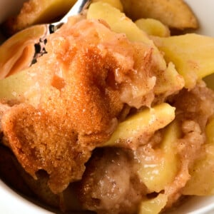 Apple cobbler featuring a cake-like topping over baked apples in a white bowl with a silver spoon.