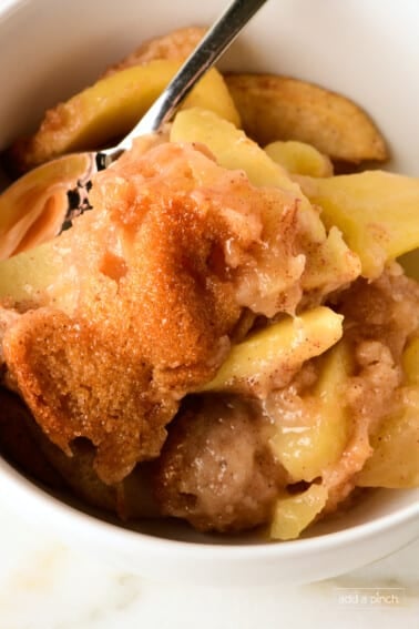 Apple cobbler featuring a cake-like topping over baked apples in a white bowl with a silver spoon.