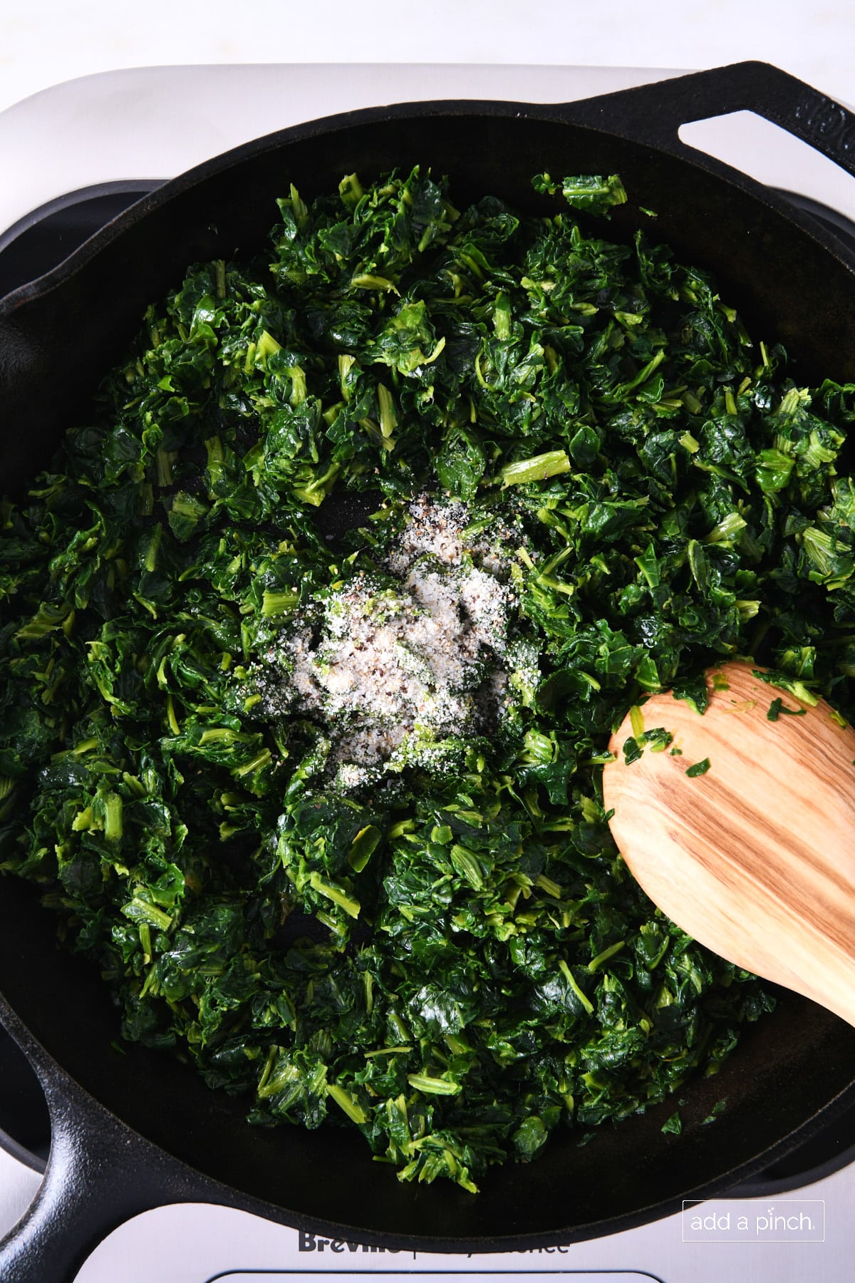Seasoning added to spinach in a skillet with a wooded spoon.