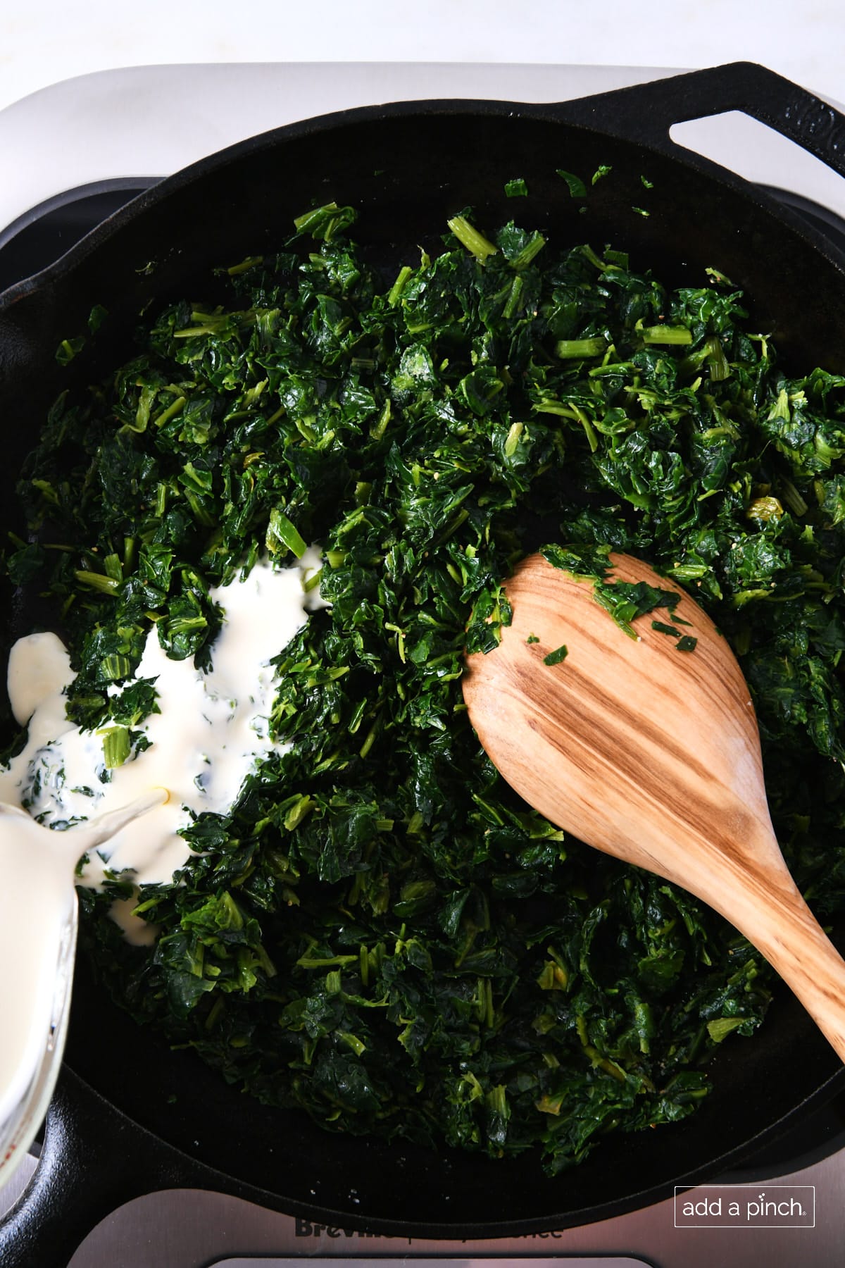 Heavy cream added to spinach in a skillet with a wooden spoon.