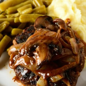 Hamburger steak topped with mushroom and onion gravy served with mashed potatoes and green beans on a white plate.