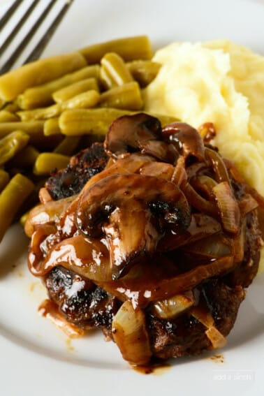 Hamburger steak topped with mushroom and onion gravy served with mashed potatoes and green beans on a white plate.