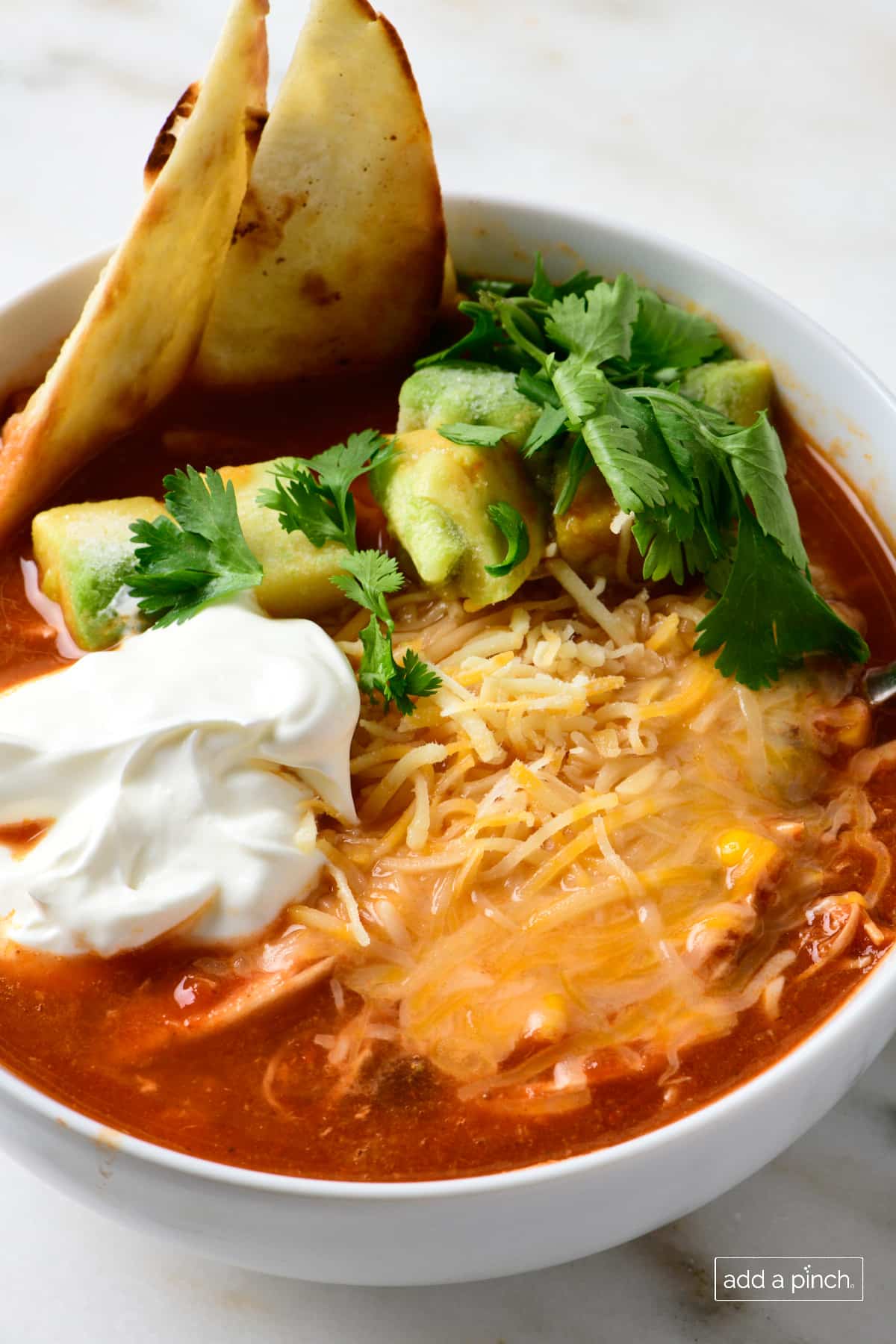 Bowl of chicken tortilla soup garnished with tortilla strips, avocados, sour cream, and shredded cheese, served in a white bowl.