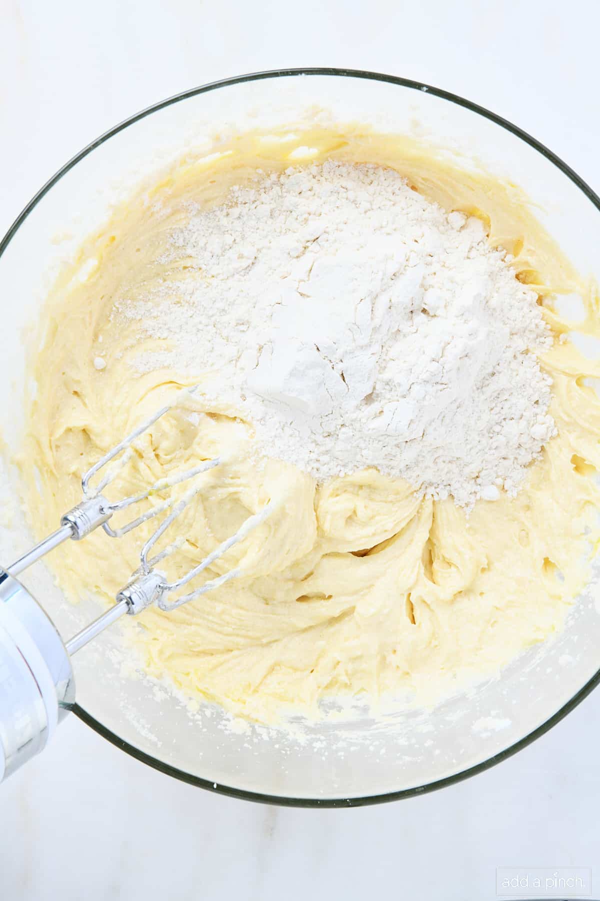 Glass mixing bowl has flour added to pound cake batter to mix with hand mixer.