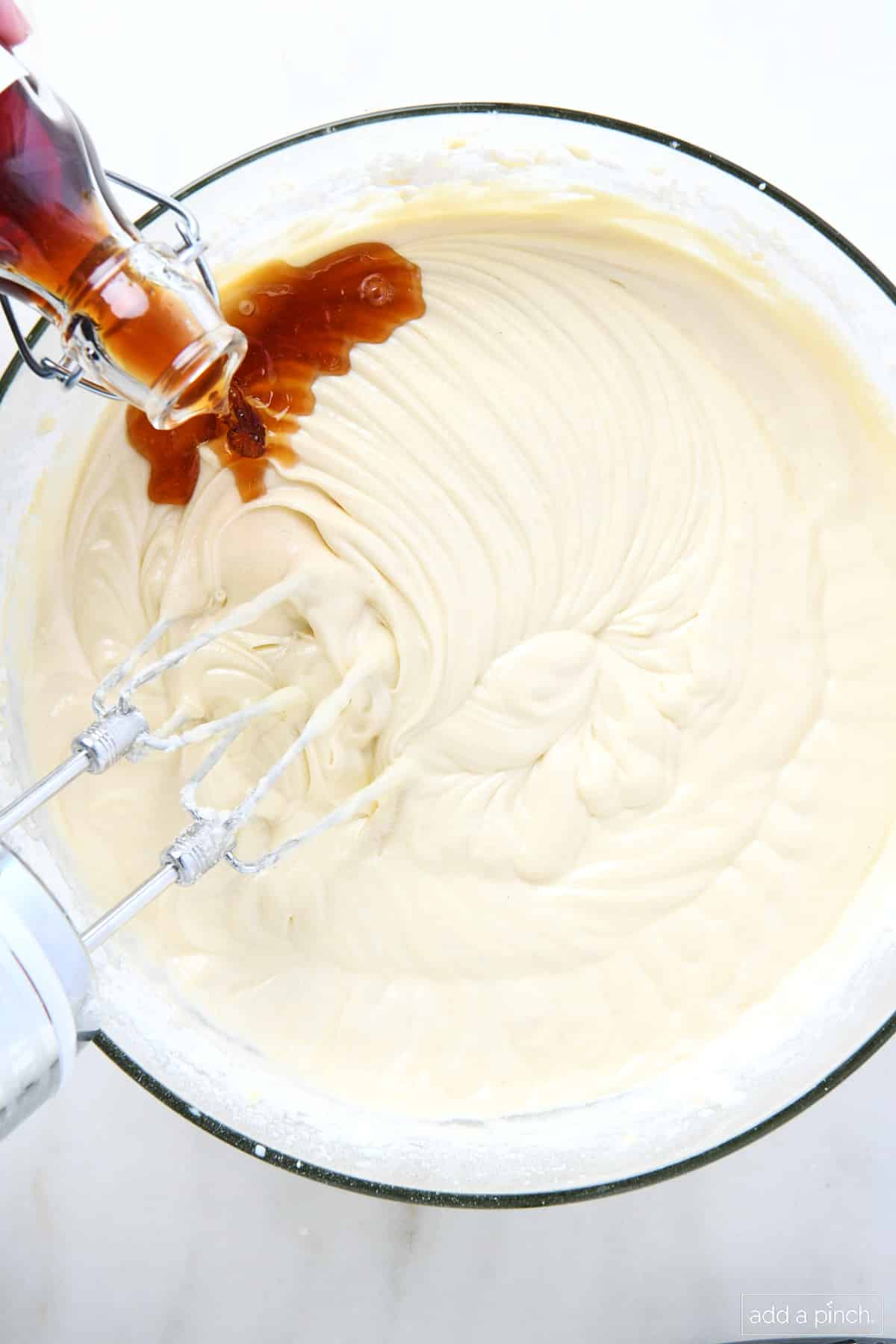 Homemade vanilla extract from a glass bottle is poured into a mixing bowl filled with pound cake batter, ready to combine with the hand mixer.