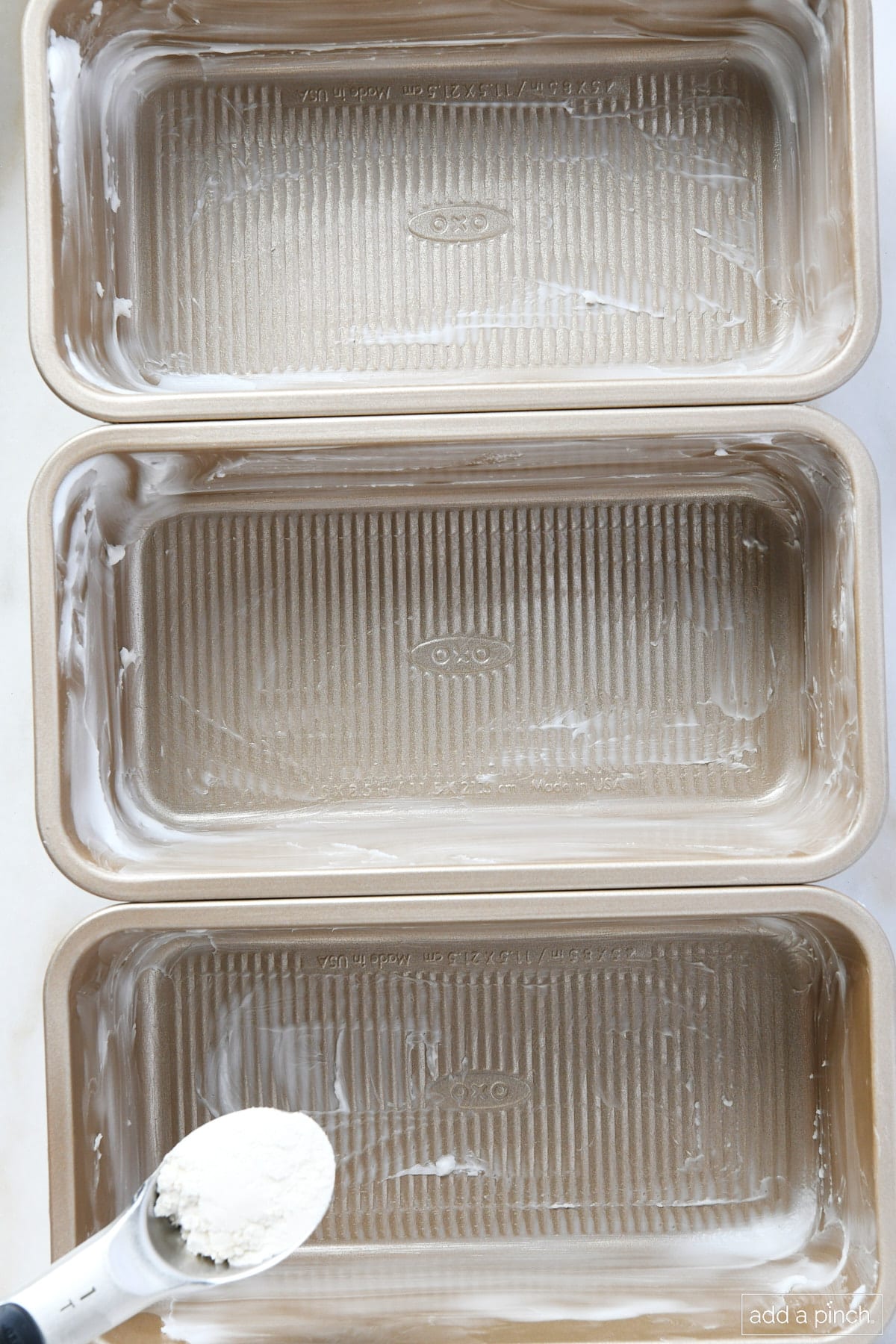 Three greased loaf pans about to have flour sprinkled inside to prep them for baking cakes.