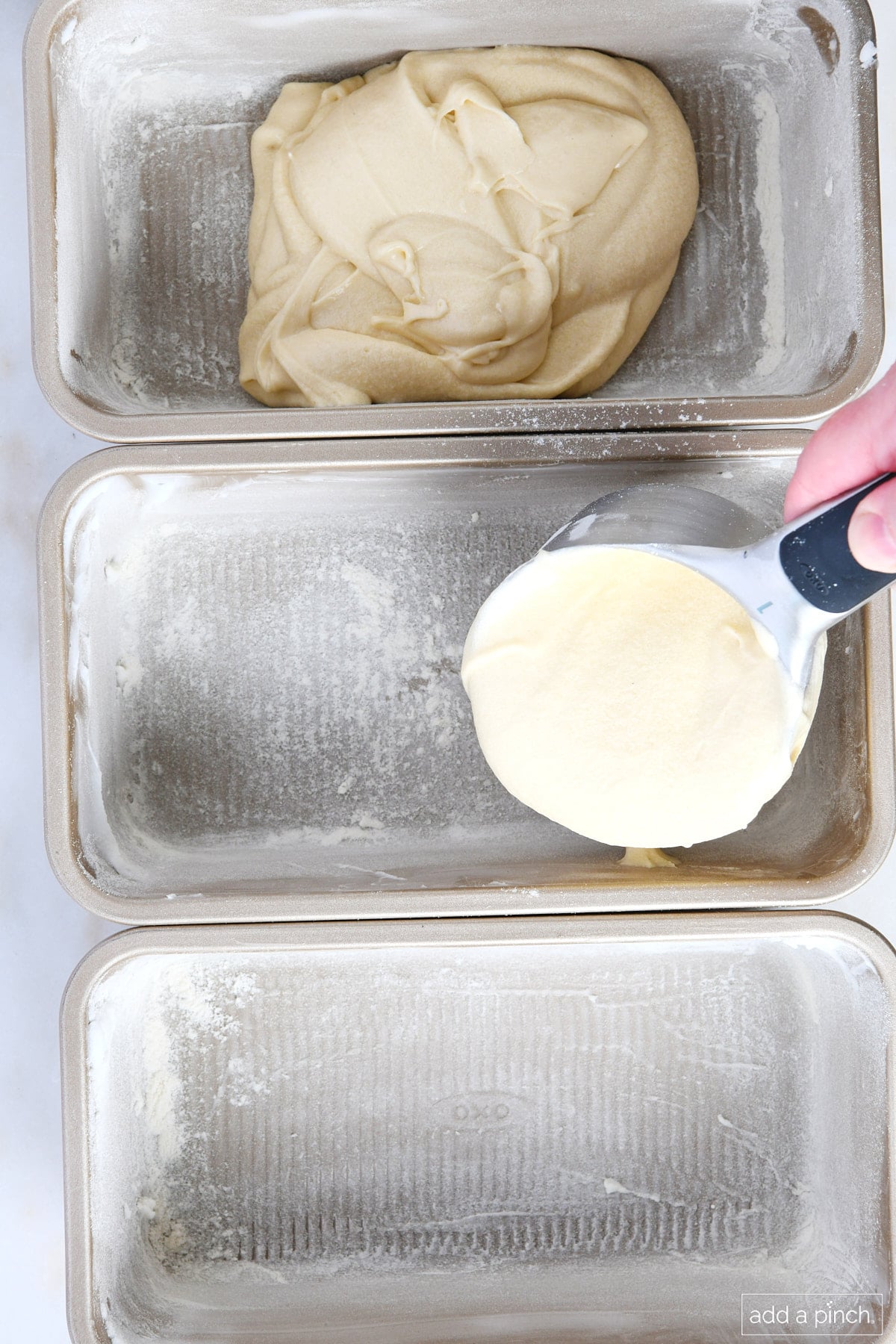 Three metal loaf pans that have been prepped with shortening and flour are having pound cake batter scooped into them. 