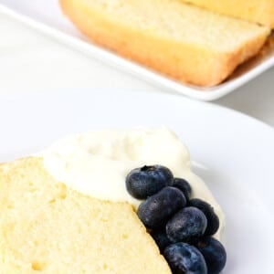 Slice of pound cake with whipped cream and fresh blueberries on a white plate, adjacent to a serving platter with sliced loaf of cake.
