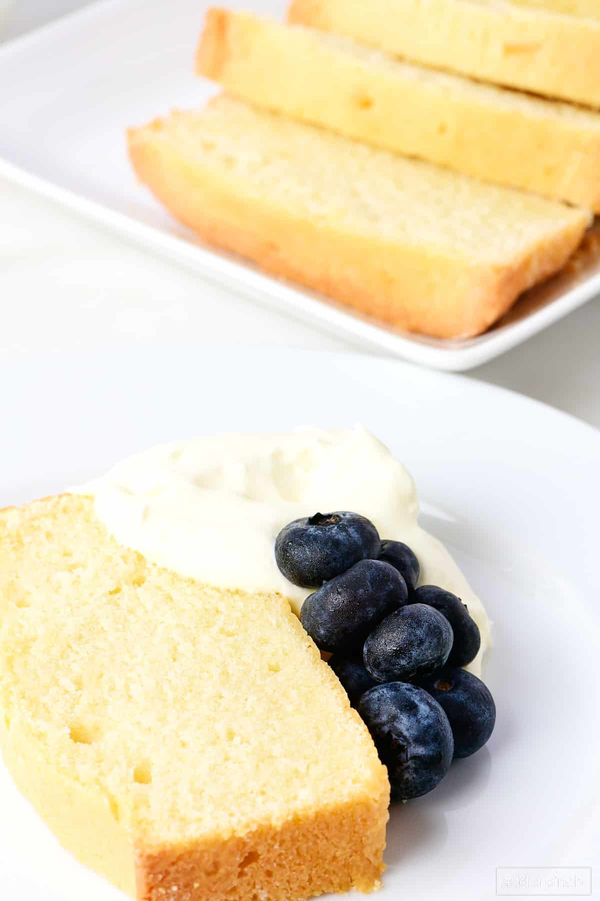 Slice of pound cake garnished with whipping cream and blueberries, with sliced cake in the background on a serving tray.